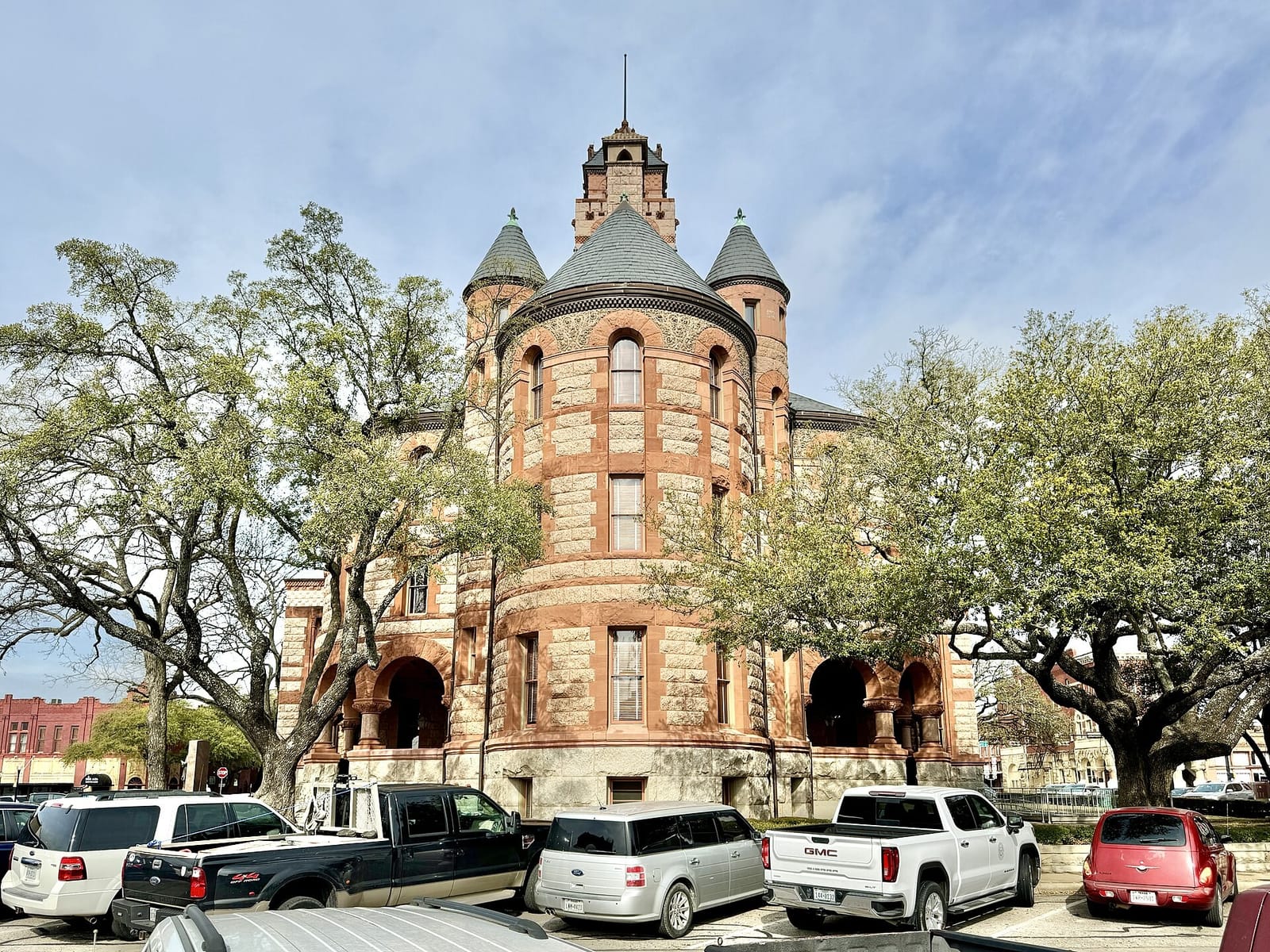 The Ellis County Courthouse in Waxahachie, Texas, a Romanesque Revival building of pink granite banded with red sandstone, with round turrets, a gray slate roof, and a stone clock tower above live oaks