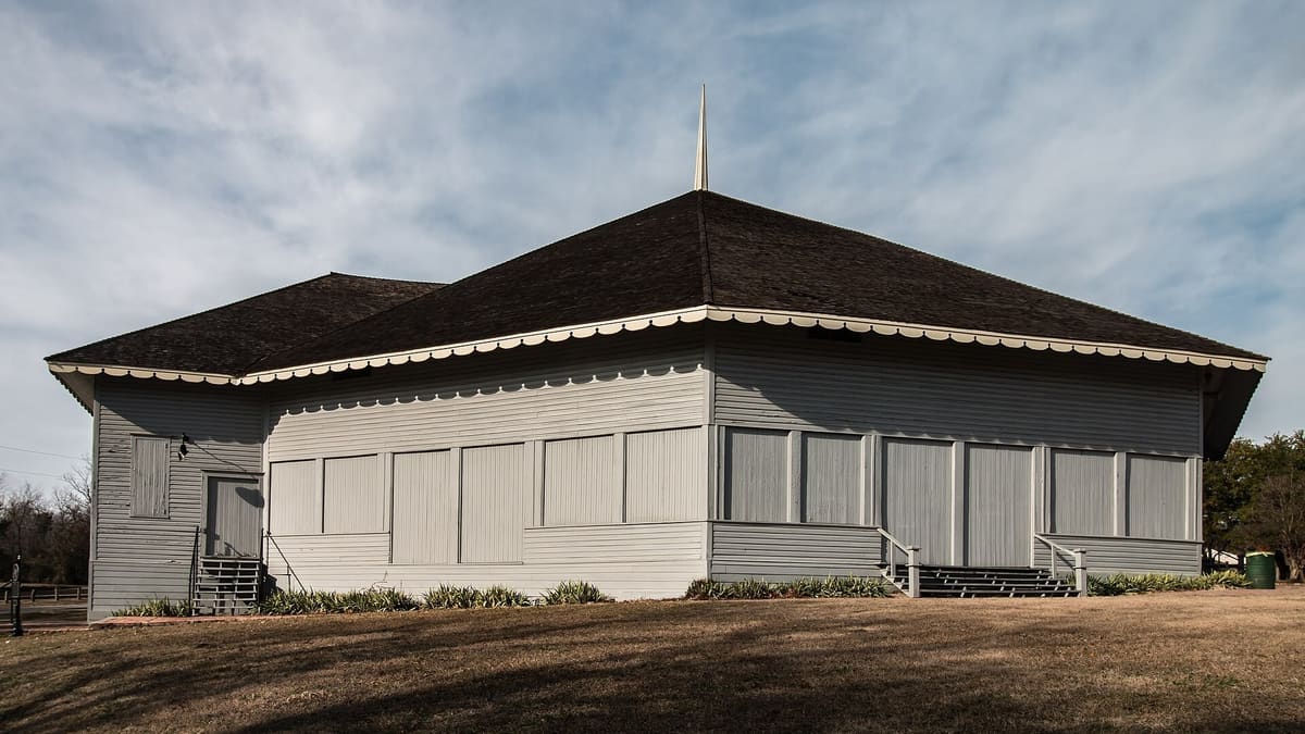 The Waxahachie Chautauqua Auditorium in Getzendaner Park, a broad gray wood building with a dark hipped roof, scalloped white eave trim, and a tall spire