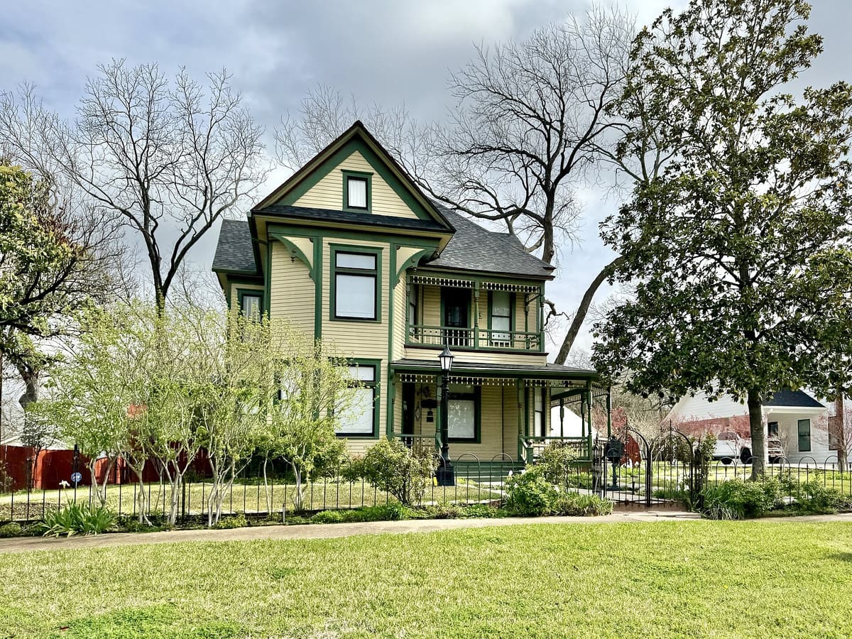 The J. M. Alderdice House on West Main Street in Waxahachie, a two-story Queen Anne house with cream siding, dark green trim, corner brackets, and a two-level spindlework porch behind an iron fence