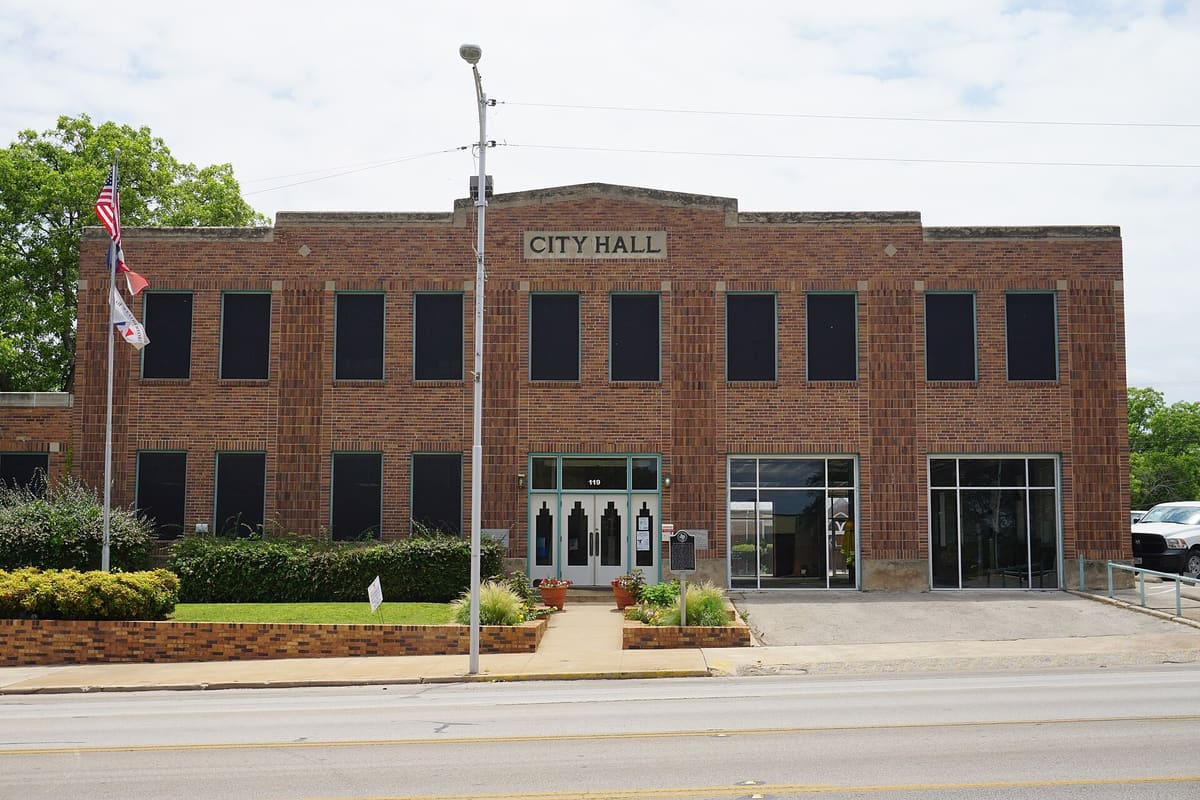 The 1933 Weatherford City Hall at 119 Palo Pinto Street, a two-story brown brick building with vertical pilasters, a stone City Hall panel, and wide glass openings at the right