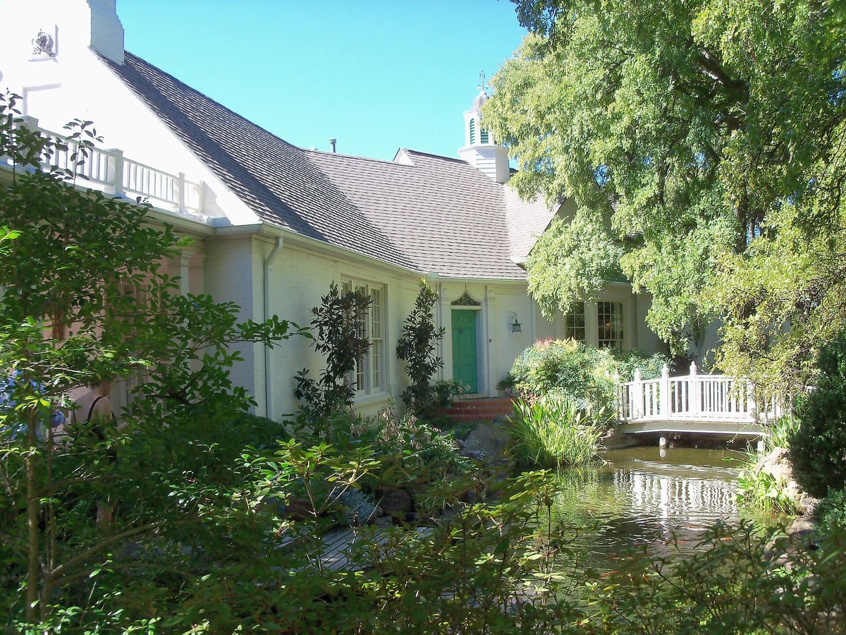 The white painted house at Chandor Gardens in Weatherford with a gray shingle roof and cupola, a green front door, and a white footbridge over a koi pond