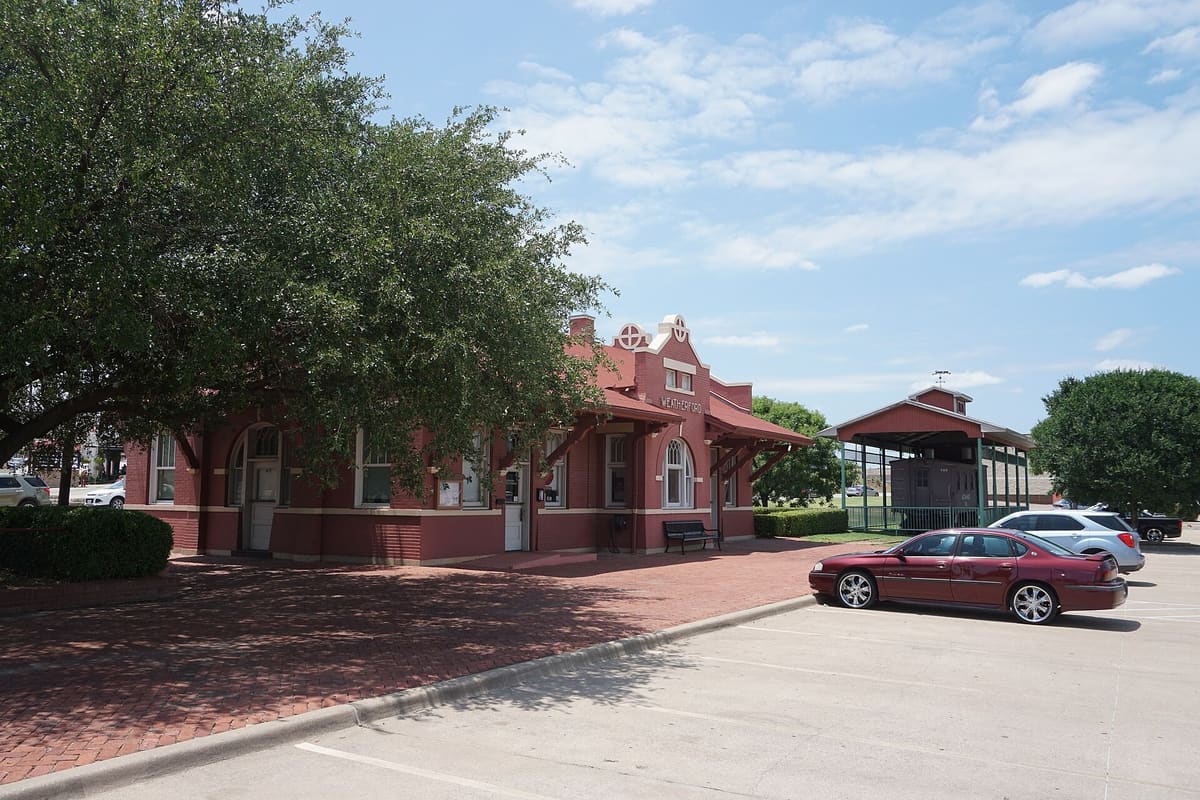 The red brick Santa Fe Depot in Weatherford with a shaped parapet reading Weatherford, an arched window, deep bracketed eaves, and a herringbone brick platform