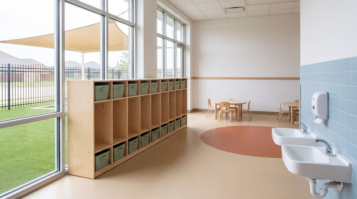 A finished toddler classroom in a newer single-story childcare center with low birch cubbies, child-height sinks, a soft rubber floor, and large windows facing a fenced play yard