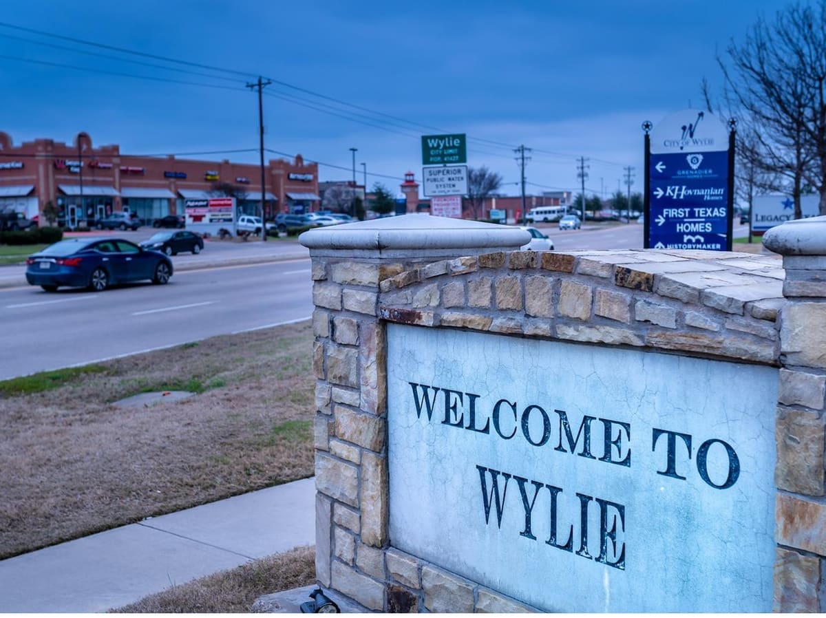 The stone Welcome to Wylie monument on FM 544 in Wylie, Texas, with a brick retail strip and traffic across the road behind it