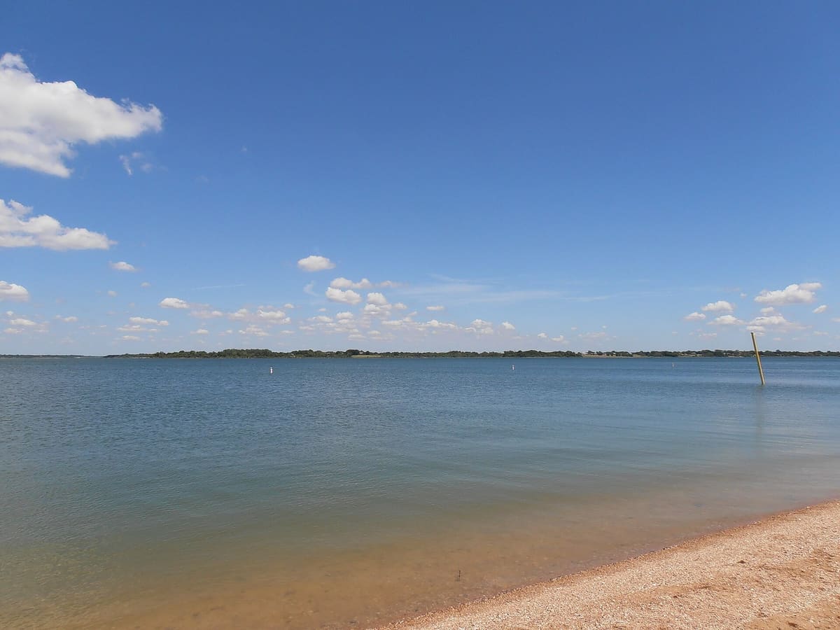 Calm blue water of Lavon Lake stretching to a low tree line under scattered clouds, seen from the sandy shore at East Fork Park in Wylie, Texas